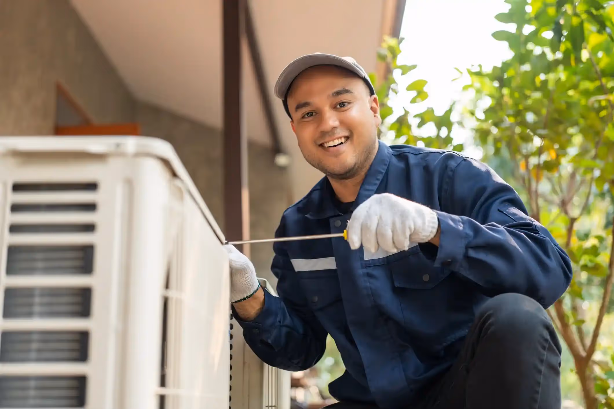 A smiling air conditioner technician is kneeling and using a screwdriver to work on an outdoor AC unit.