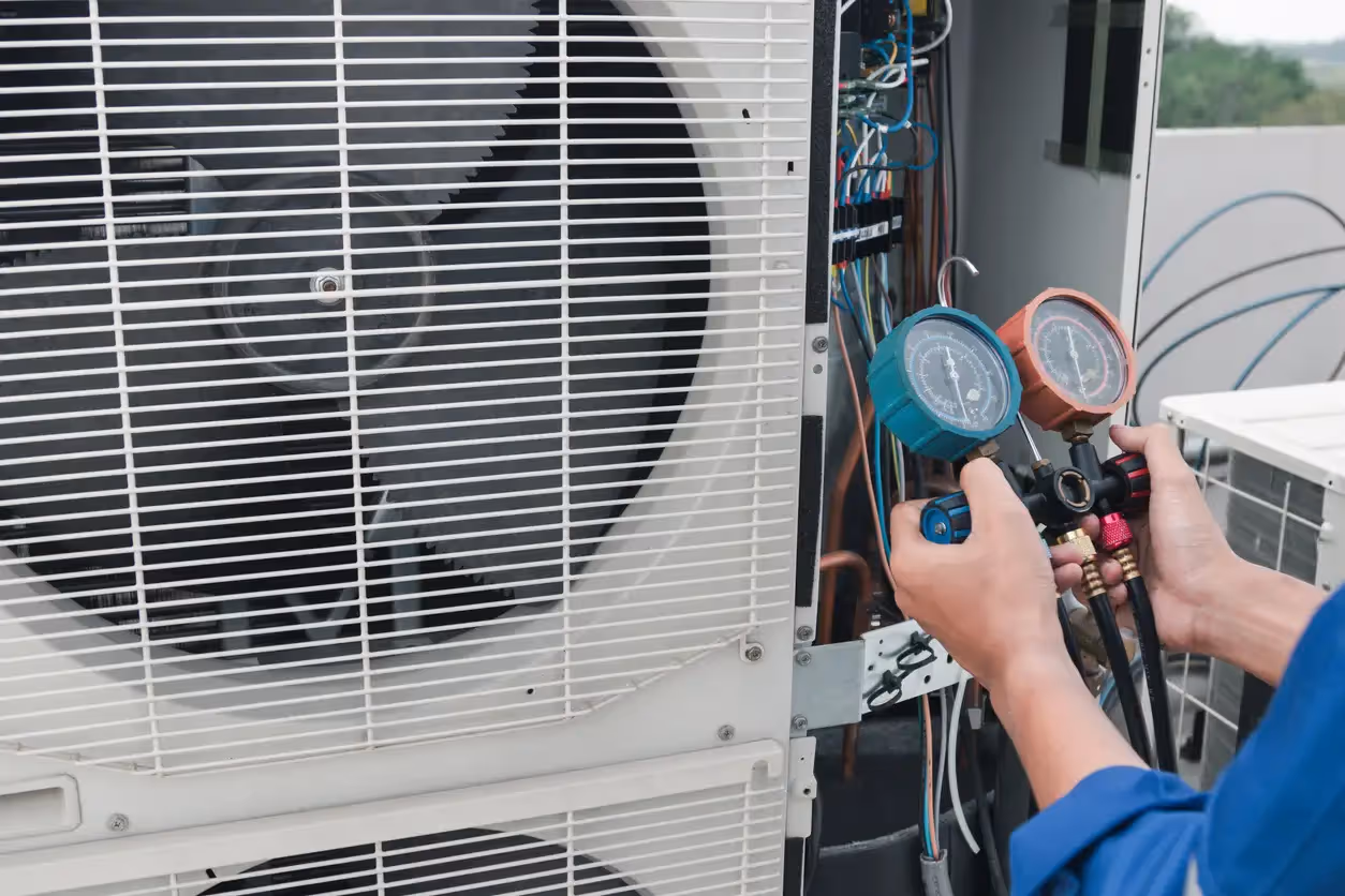 Technician holds manifold gauge set with blue and red dials connected to open HVAC unit showing internal components.