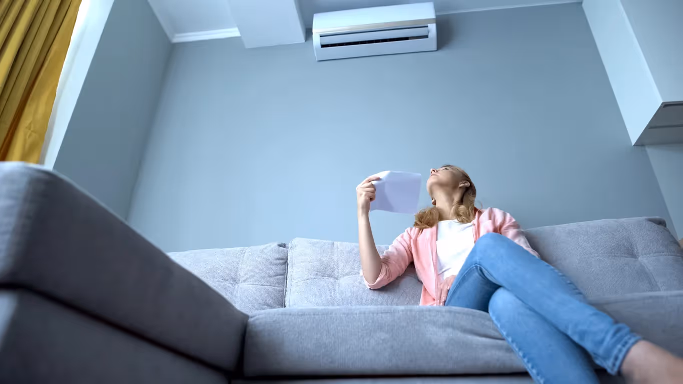 A woman fanning herself on a couch under a non-working air conditioner.