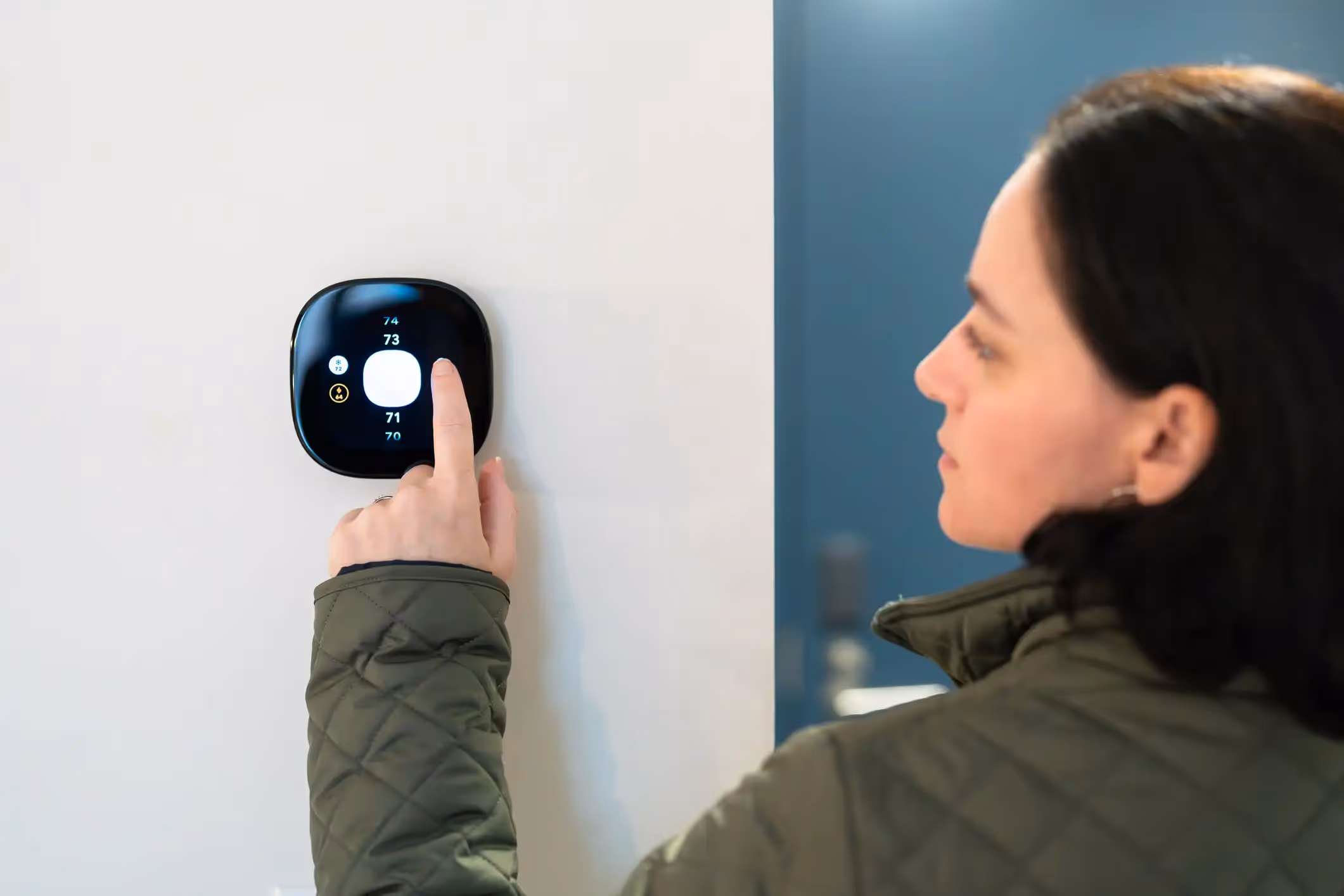 Woman pointing at a circular, black smart thermostat mounted on a white wall.