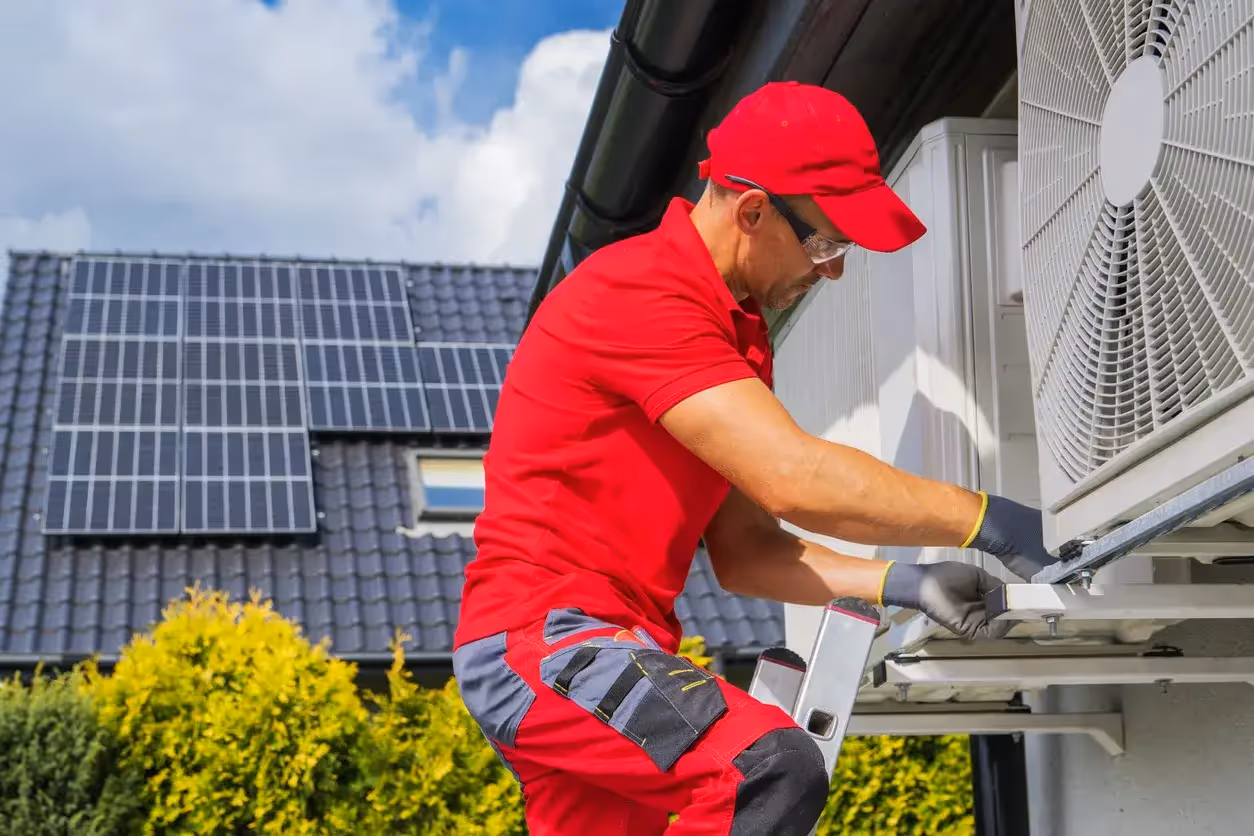 Professional installation of an outdoor heat pump unit with residential solar panels in the background.