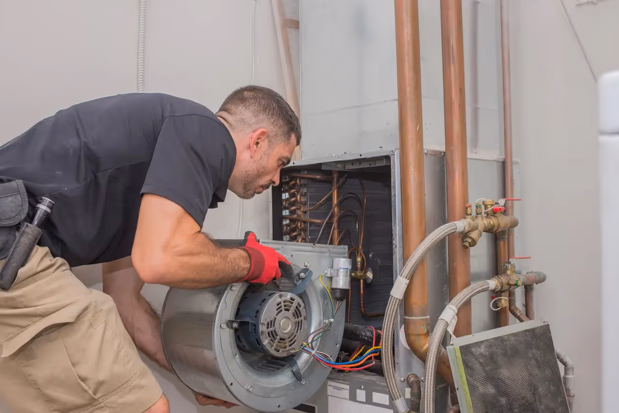 Technician in red gloves handles blower motor assembly with exposed copper pipes and coil in HVAC unit.