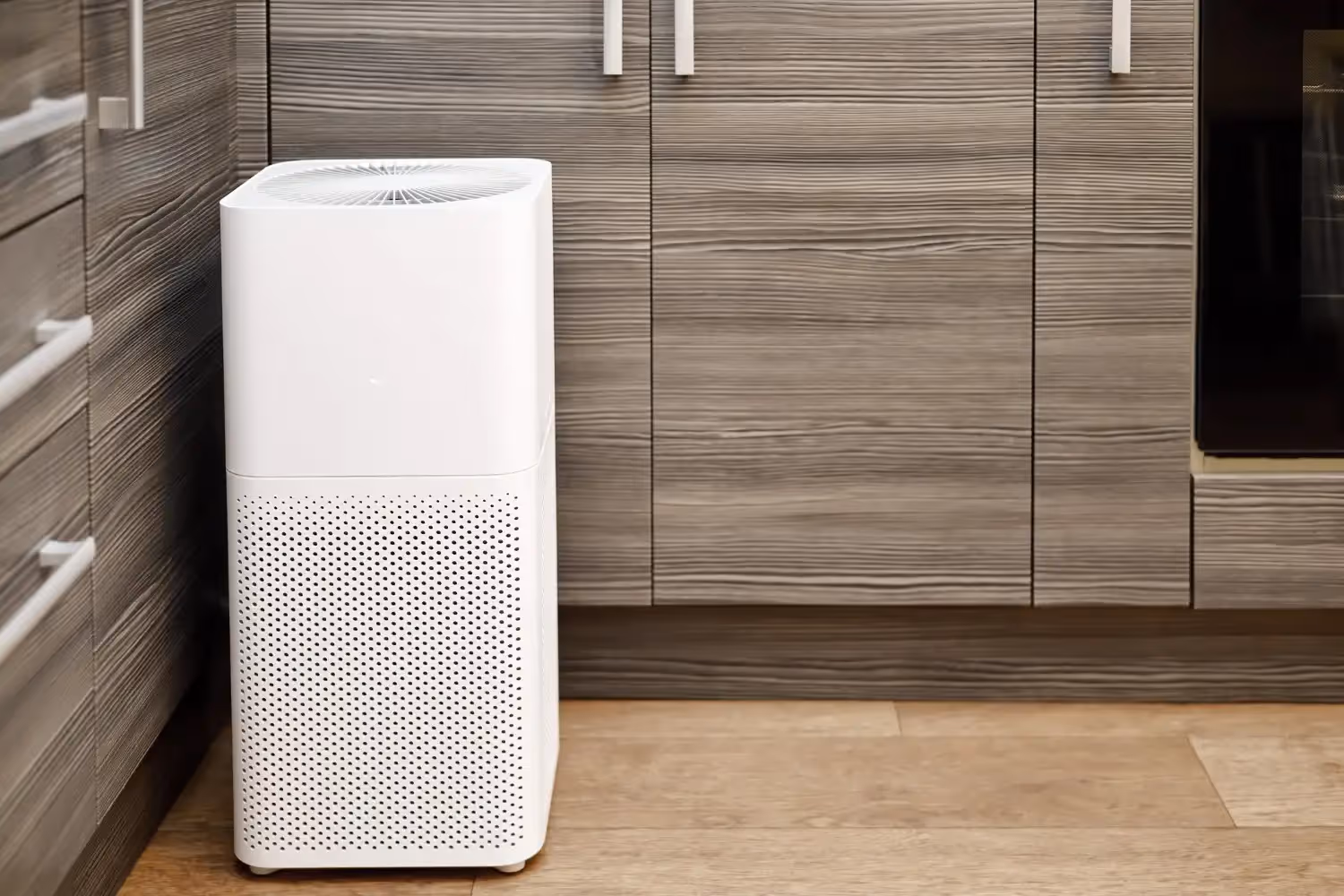 A white tower air purifier with a perforated intake base stands on a light wood floor in a modern kitchen. It is positioned against contemporary grey wood-grain cabinets.