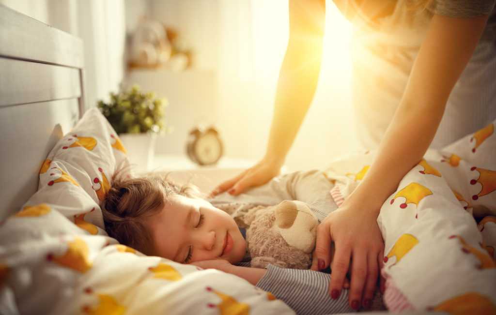 Sleeping child in bed with a teddy bear, cozy bedding, and a caregiver gently tucking them in, emphasizing comfort and care in a warm, inviting room.
