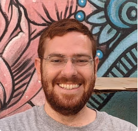 Smiling man with glasses and a beard in front of a colorful mural with pink and blue patterns.