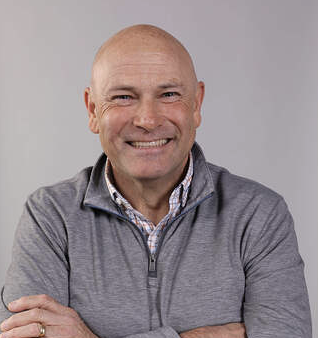 Smiling bald man wearing a grey quarter-zip sweater and checkered shirt against a light grey background.