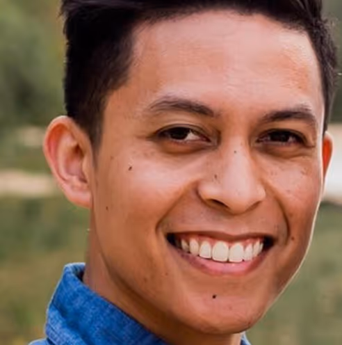 Close-up of a smiling young man with short dark hair wearing a blue shirt in an outdoor setting.