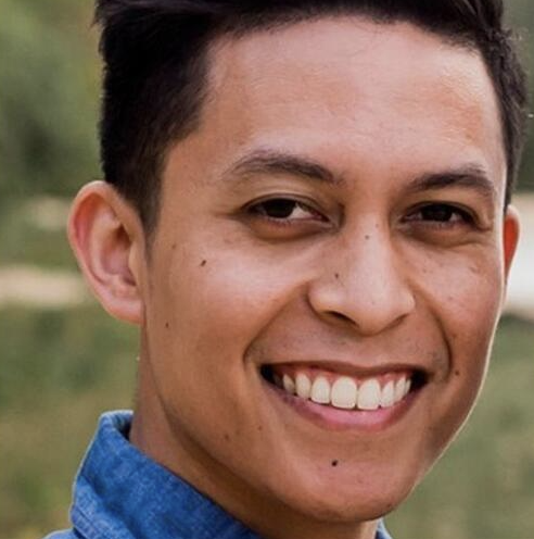 Close-up of a smiling young man with short dark hair wearing a blue shirt in an outdoor setting.