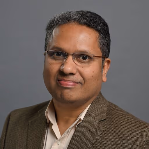 Man with short black and gray hair wearing glasses, a brown blazer, and a beige shirt against a gray background.