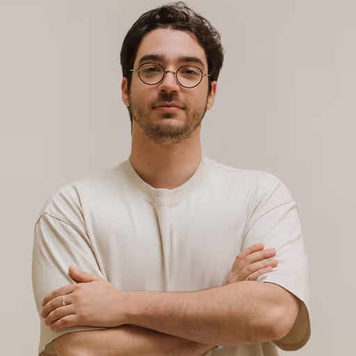 Young man with dark curly hair and glasses wearing a white t-shirt with arms crossed against a plain background.