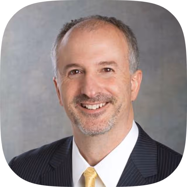 Smiling middle-aged man with short gray hair and beard wearing a dark pinstripe suit and yellow tie.
