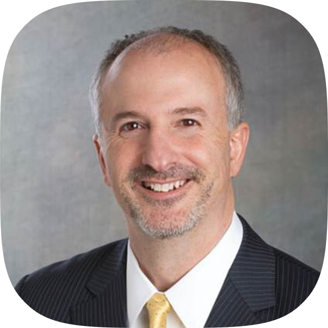 Smiling middle-aged man with short gray hair and beard wearing a dark pinstripe suit and yellow tie.