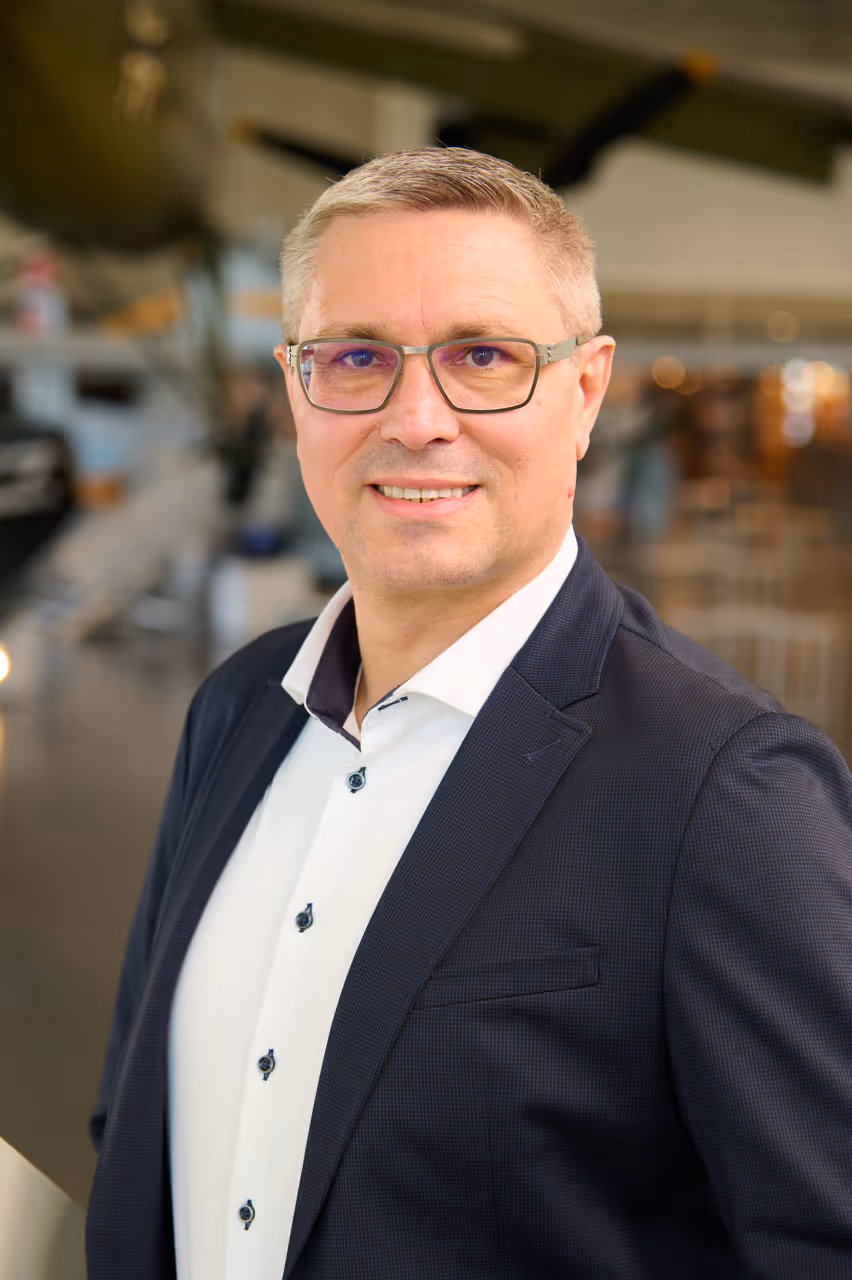 Smiling middle-aged man with glasses wearing a dark blazer and white shirt in an indoor setting.