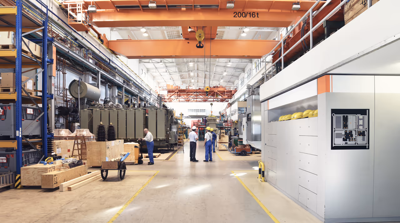 Three workers wearing helmets and overalls standing inside a large, organized industrial warehouse with machinery and equipment.