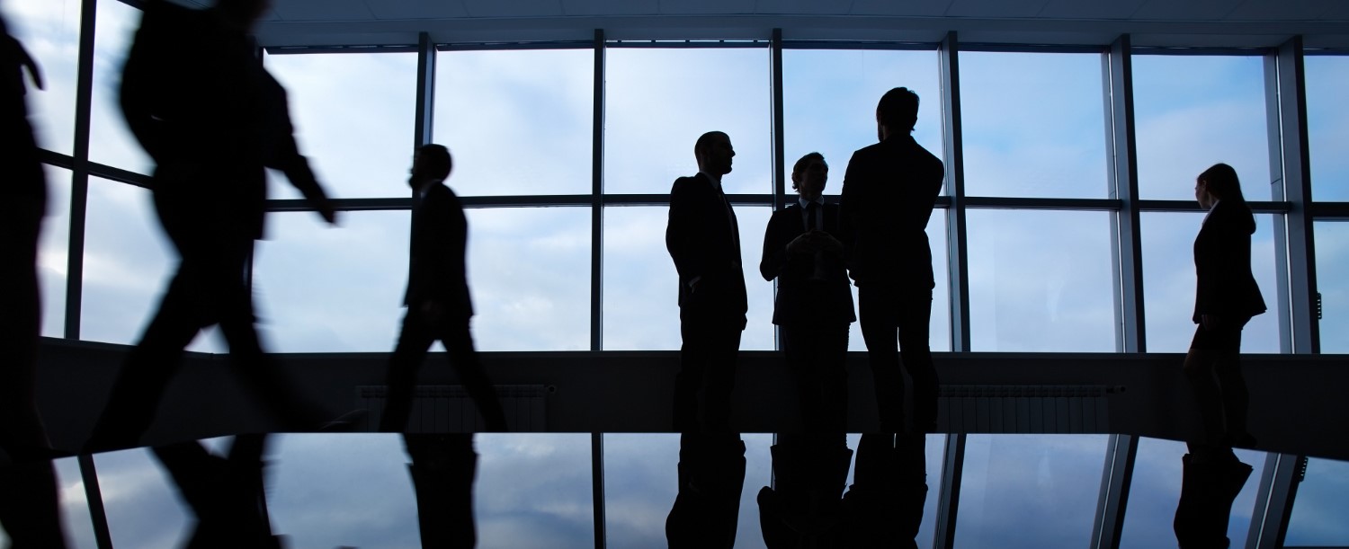 Silhouettes of business people standing and walking in an office with large windows and reflective table surface.