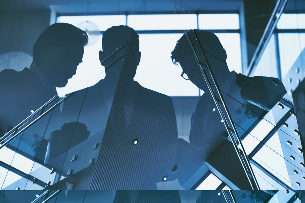Silhouettes of three businesspeople in discussion inside an office building with glass walls and a blue tint.