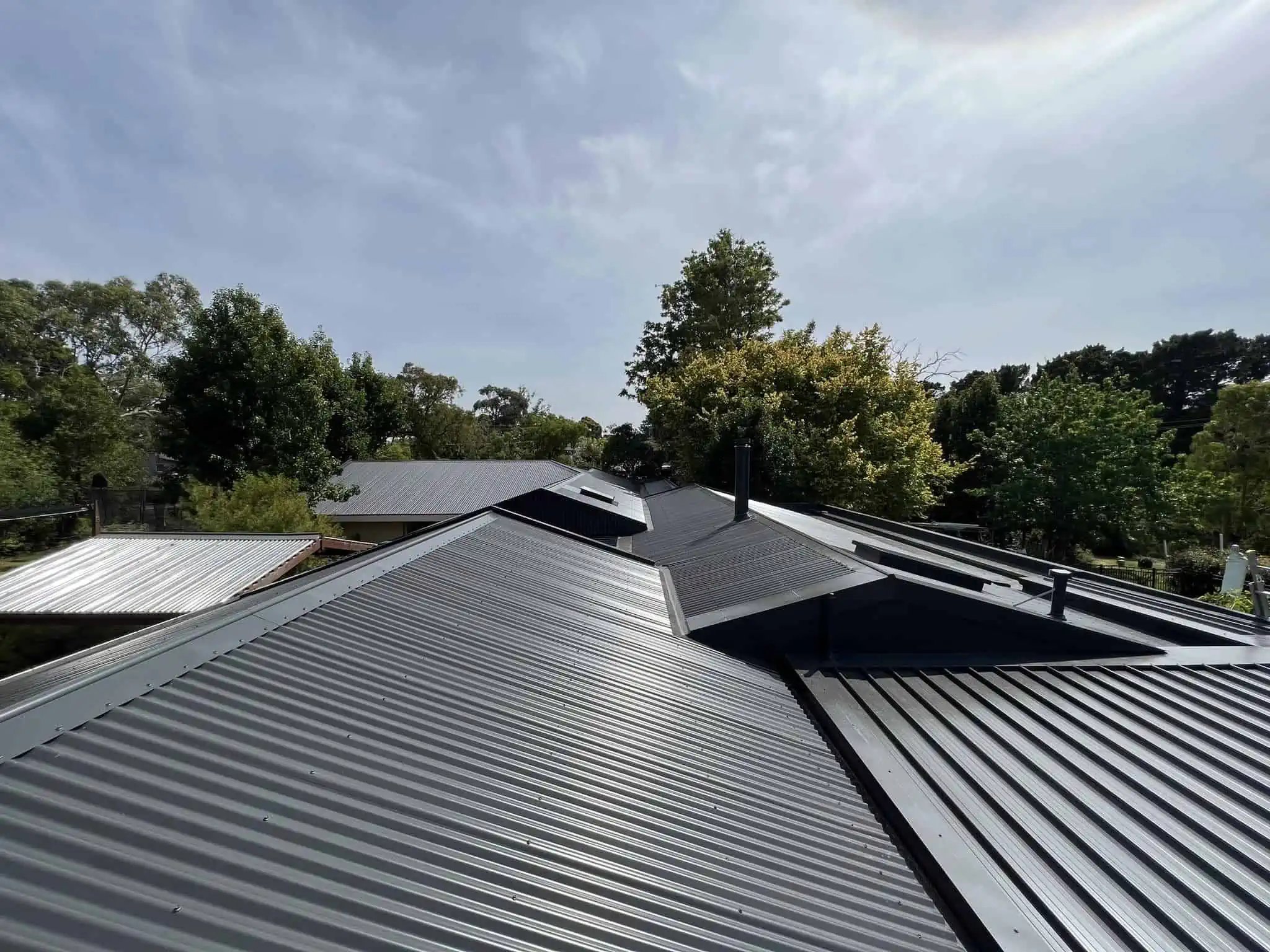 View of multiple dark gray metal roofs with trees and a cloudy sky in the background.
