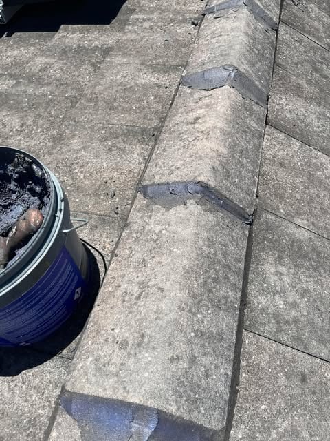 Close-up of a rooftop with concrete ridge tiles partially repaired with grey mortar and a bucket of mortar placed on the roof.