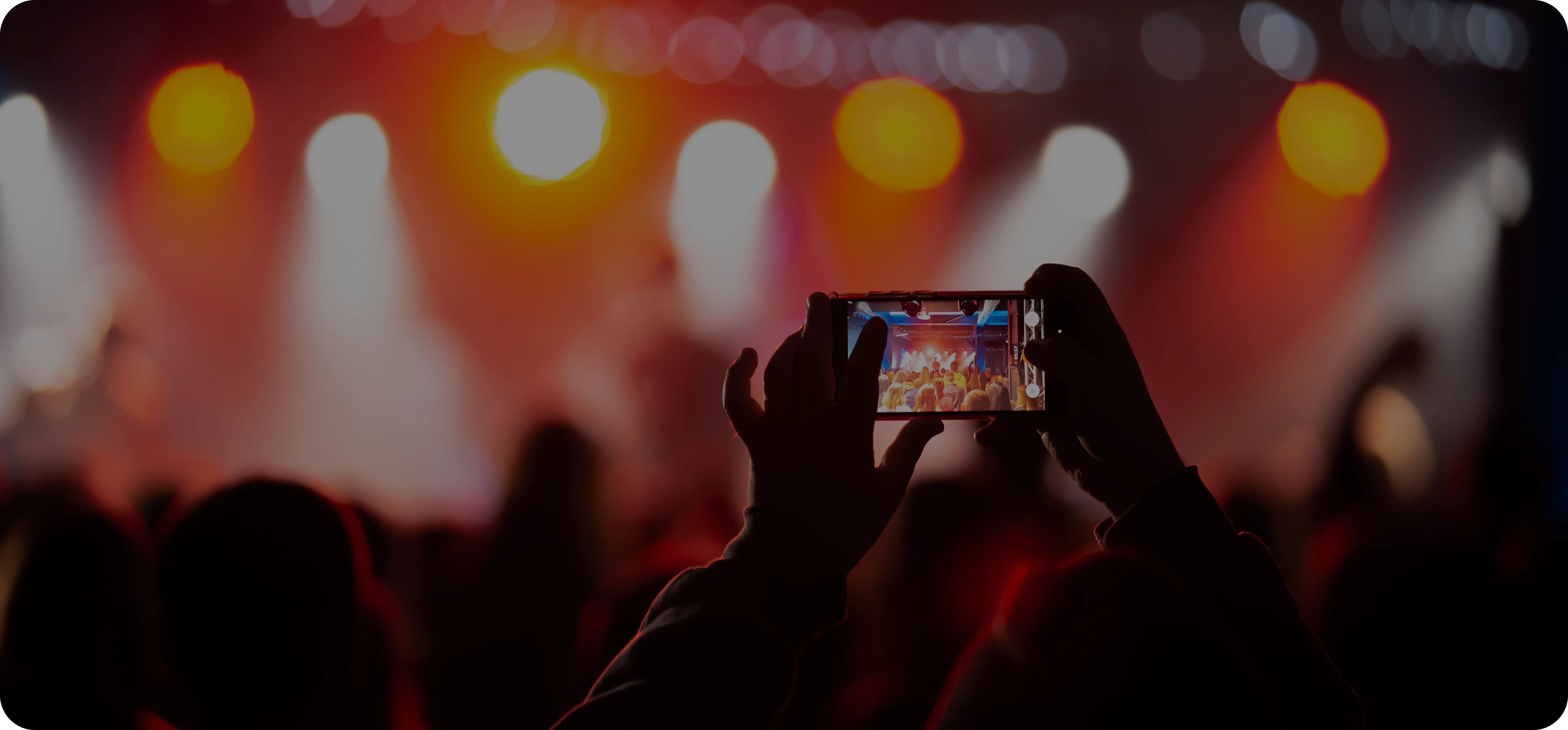 Крупний план людини, яка знімає концерт на смартфон, з яскравими сценічними вогнями та динамічною атмосферою на задньому плані.
