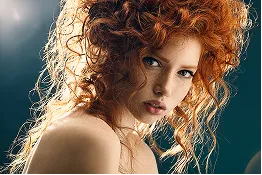 Close-up portrait of a woman with curly red hair and light skin against a dark teal background.