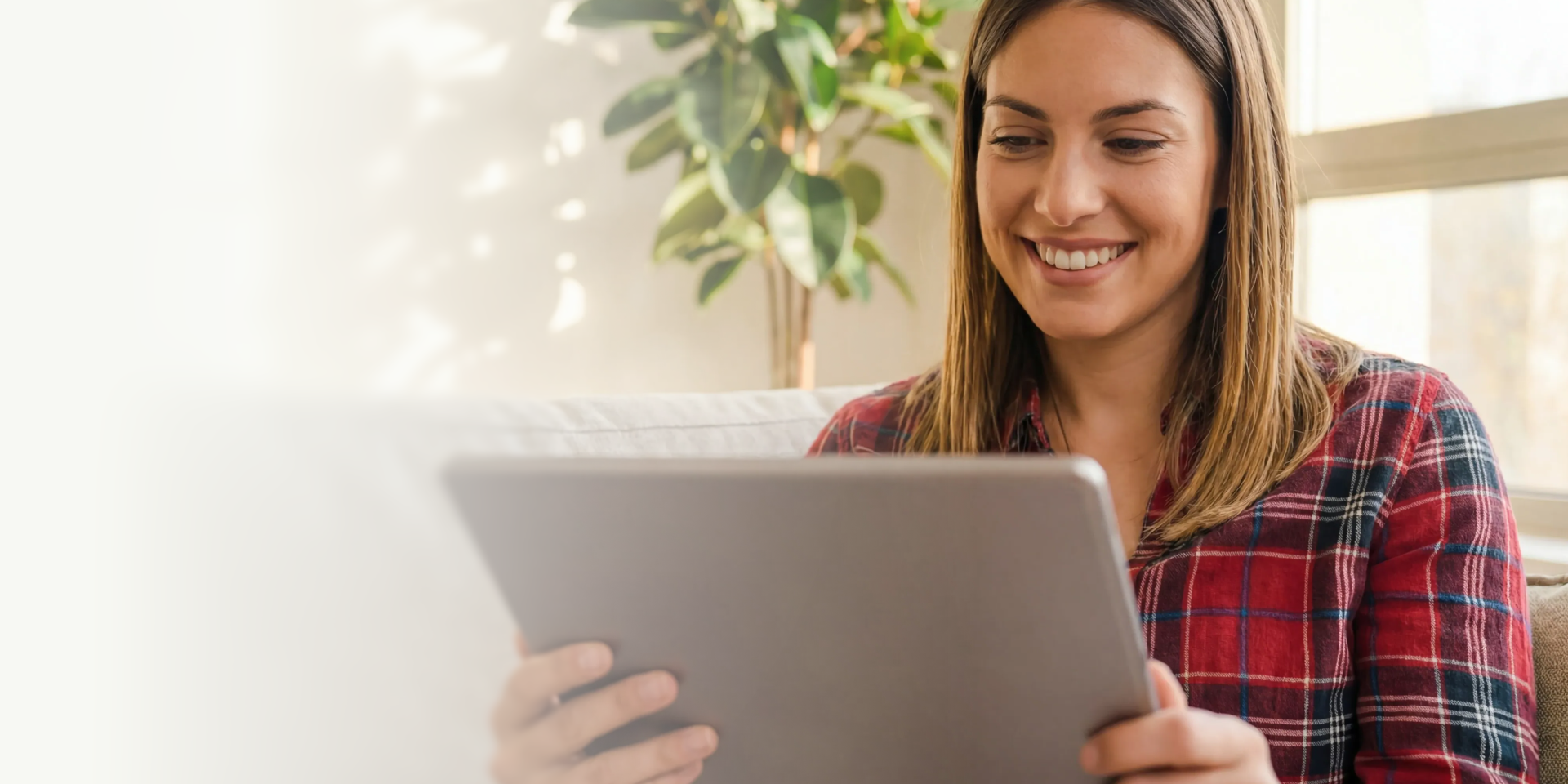 Smiling woman in a red plaid shirt holding and looking at a tablet while sitting on a couch with a plant in the background.