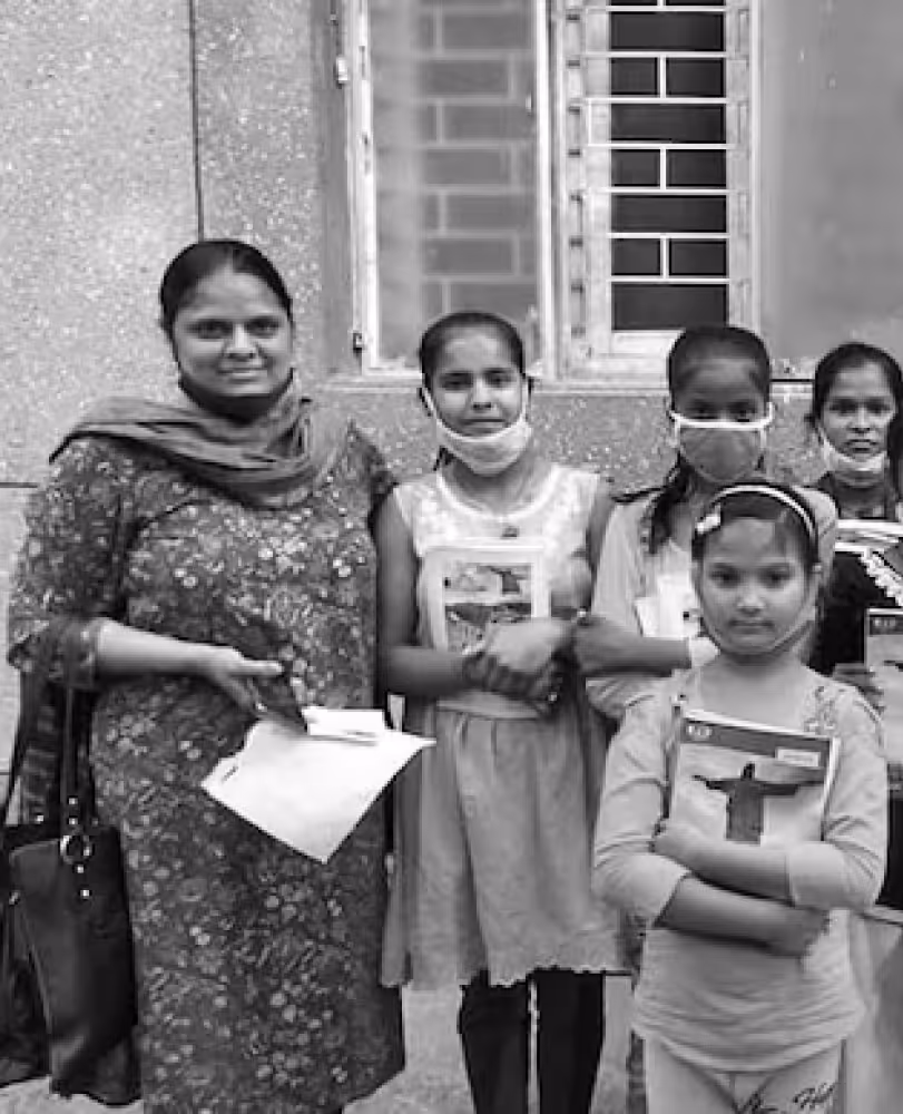 Group of smiling Indian woman and four girls, some wearing face masks, standing outdoors holding books and papers.