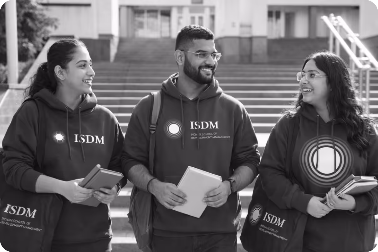 Three students wearing ISDM hoodies and carrying books and bags, smiling and walking outside near stairs.