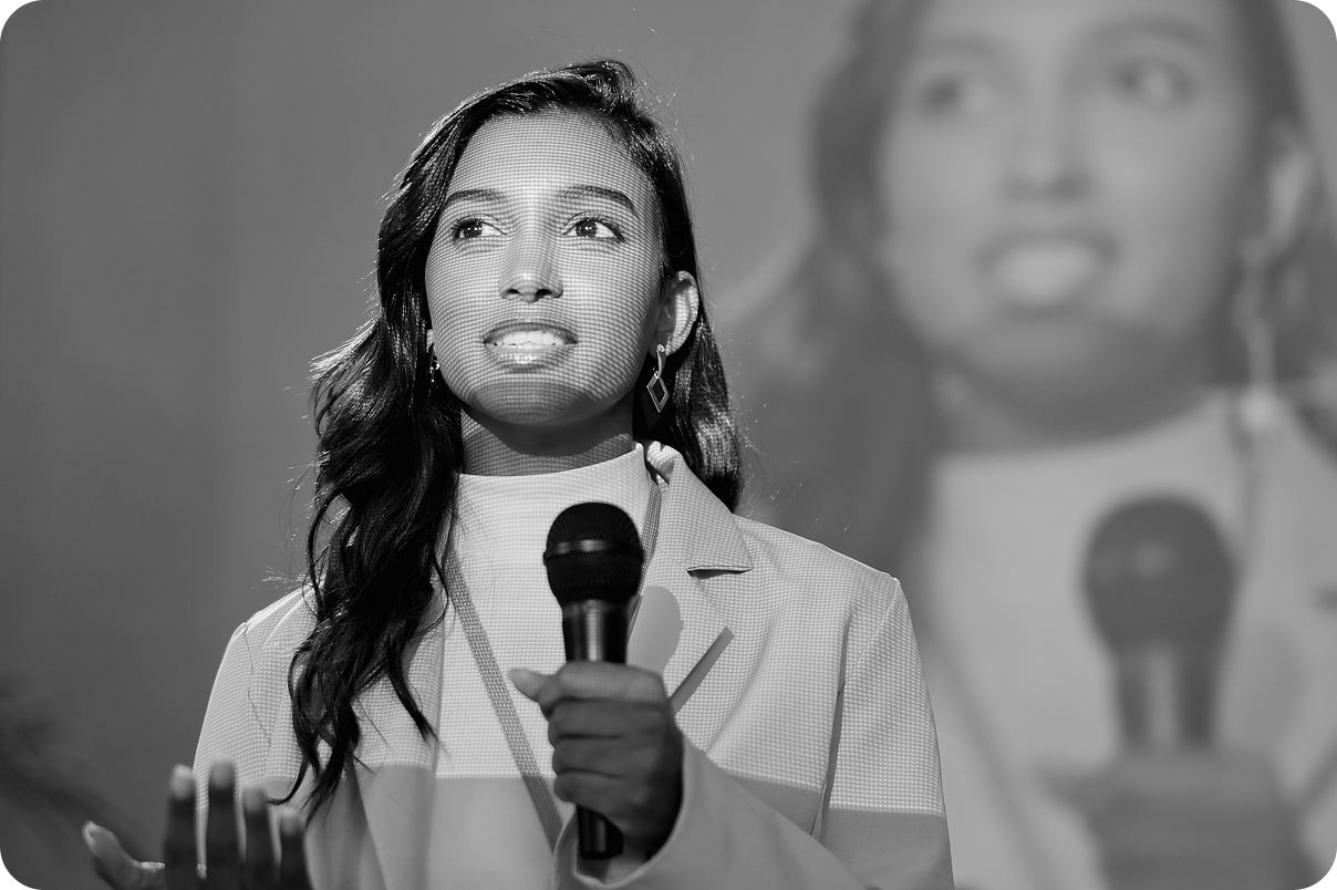 Young woman with long hair holding a microphone and speaking, with her image projected on a screen behind her.