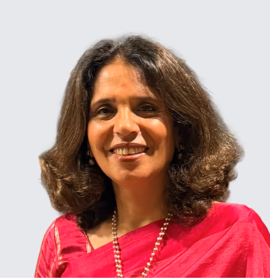 Smiling woman with wavy dark hair wearing a red saree and a pearl necklace against a light background.