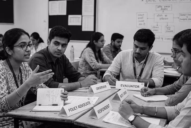 Young people seated around a table in discussion with role name tags like Policy Advocate and Evaluator.