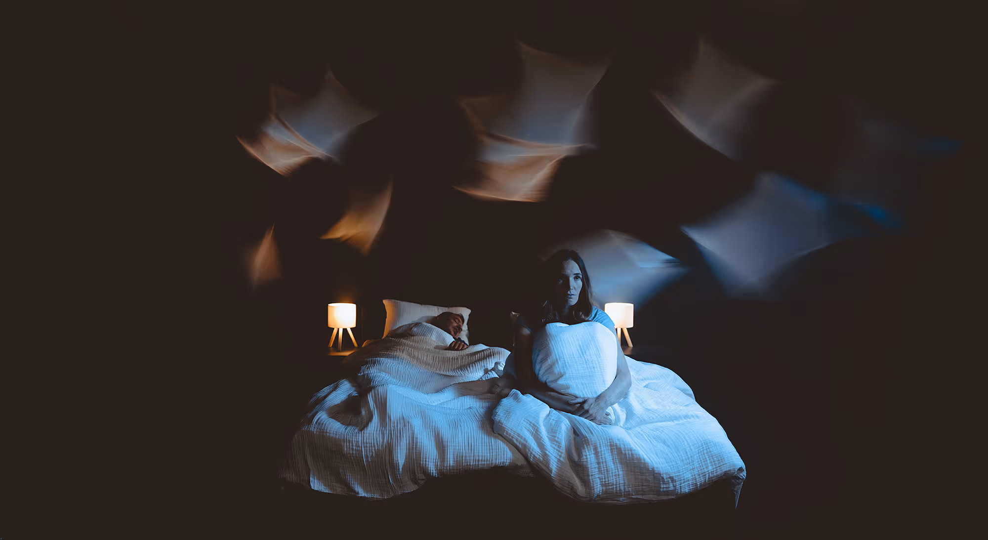 Frau sitzt nachts wach im Bett und drückt ein Kissen, während ein Mann neben ihr schläft.