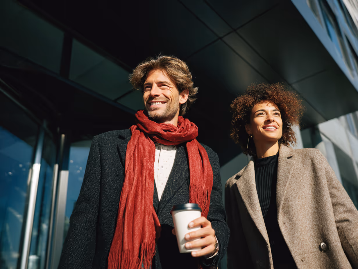 Smiling man and woman wearing coats, the man holds a coffee cup outdoors in daytime.
