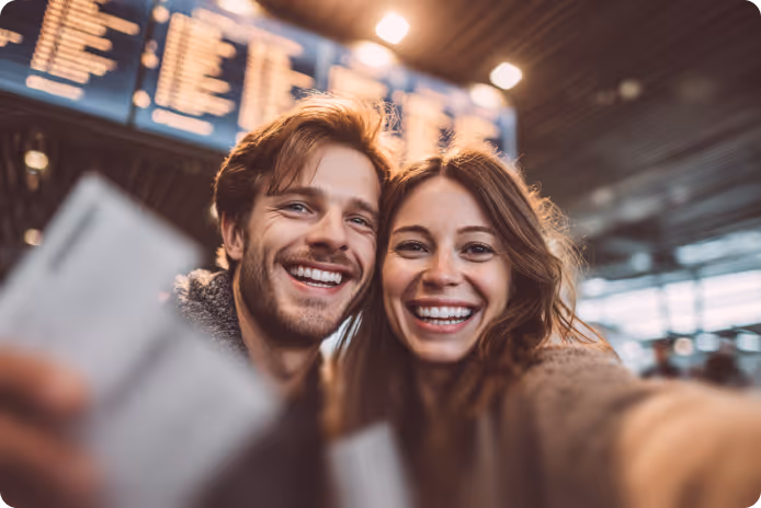 Smiling young couple holding plane tickets and taking a selfie at an airport.