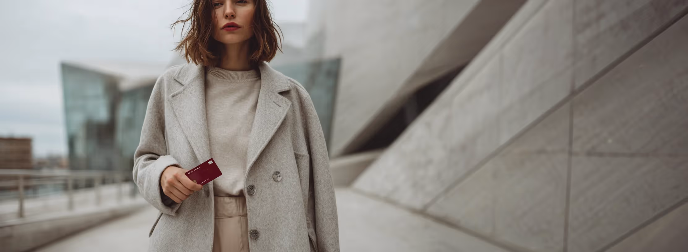 Woman wearing a light gray coat holding a red credit card outdoors in an urban setting.