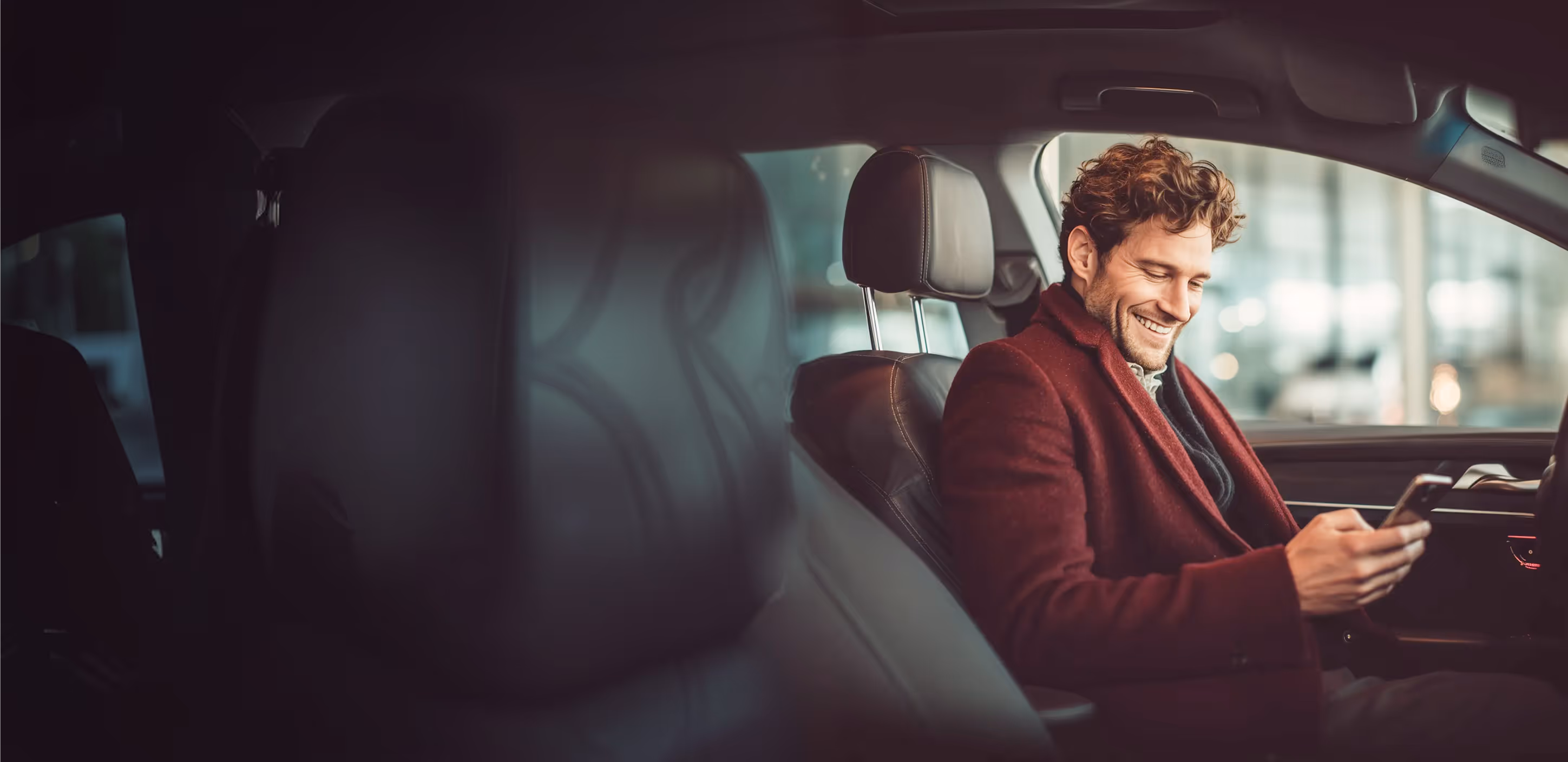Smiling man in a red coat sitting in a car looking at his smartphone.