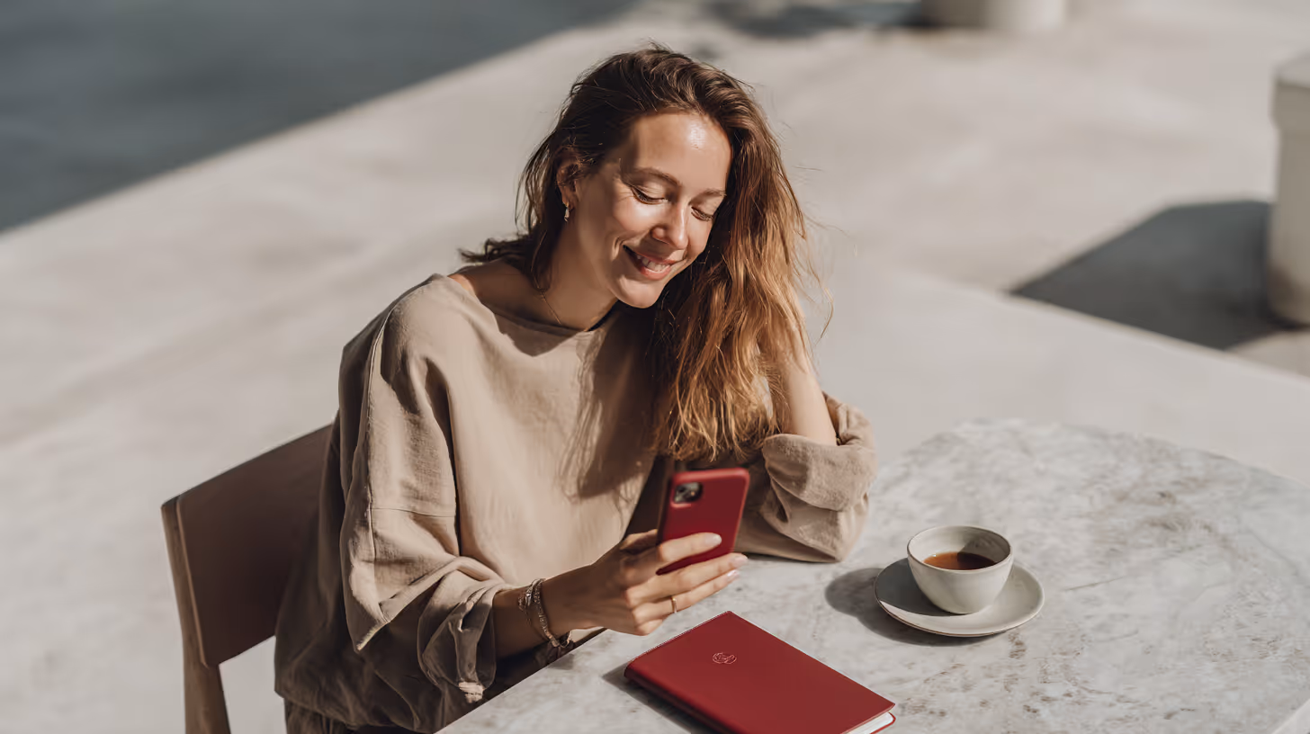 Woman smiling and looking at her phone while sitting at a marble table with a cup of coffee and a red notebook.