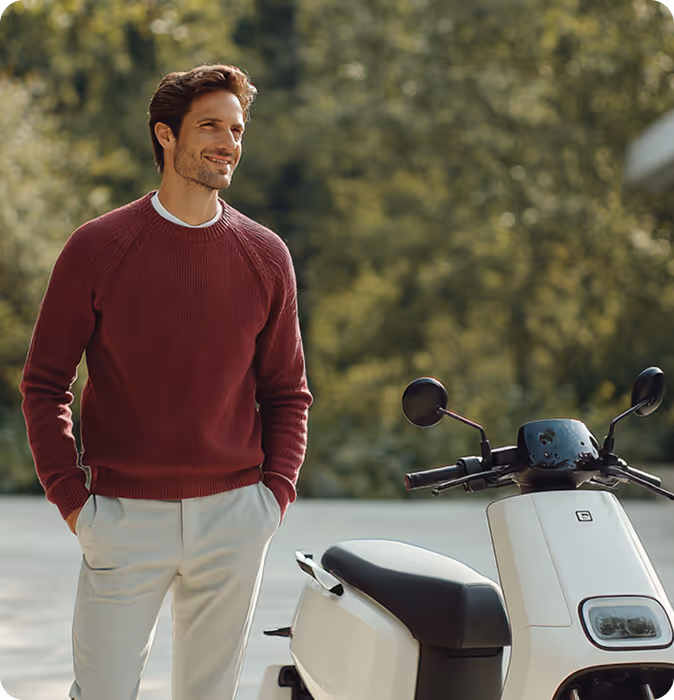 Mand iført rød trøje og beige bukser står ved siden af en hvid scooter udendørs.