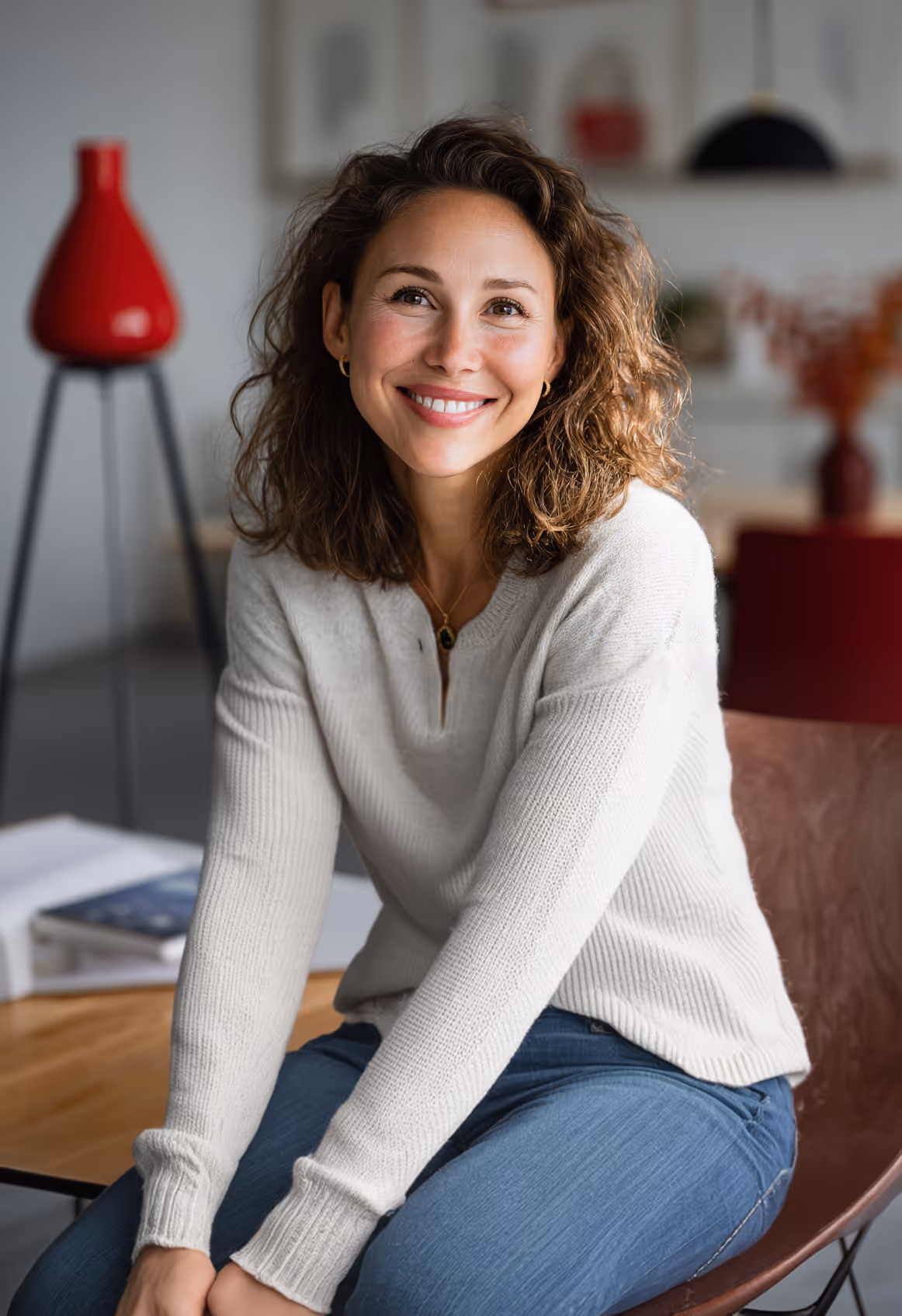 Smiling woman with curly hair wearing a white sweater and blue jeans sitting indoors.