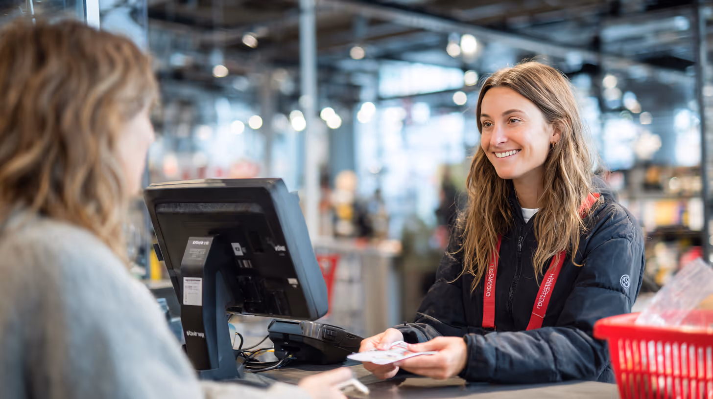 En smilende kvindelig kassemedarbejder i samtale med kunde ved kassen i et butiksmiljø.