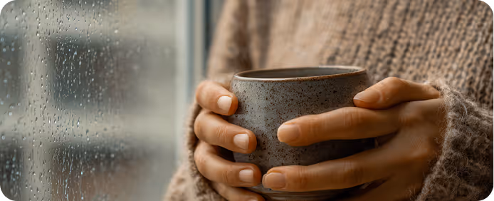 Person in a cozy brown sweater holding a speckled ceramic mug near a rainy window.