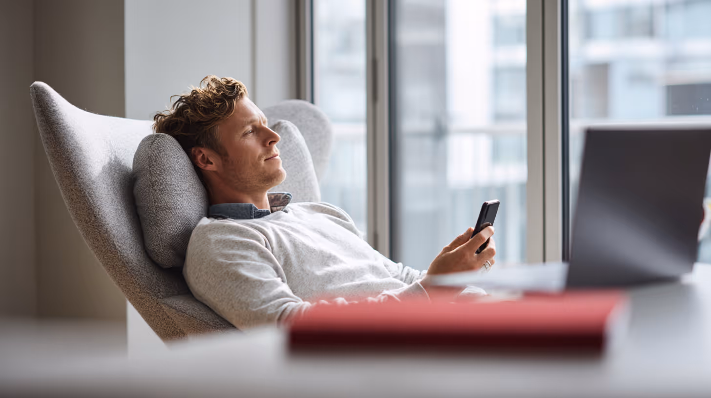 Man reclining in a gray armchair holding a smartphone next to a laptop on a desk by the window.