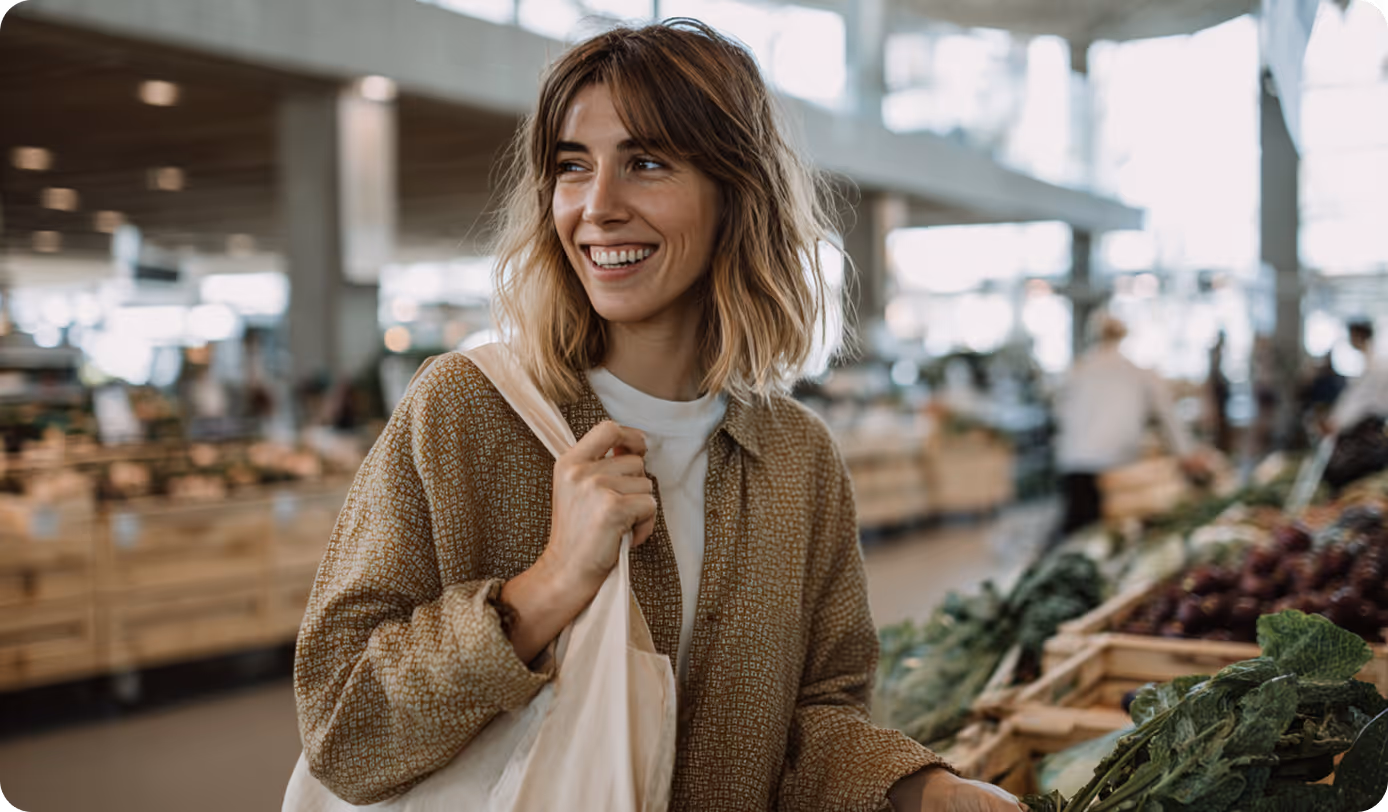 Smiling woman shopping for fresh vegetables at a market with reusable tote bag over her shoulder.