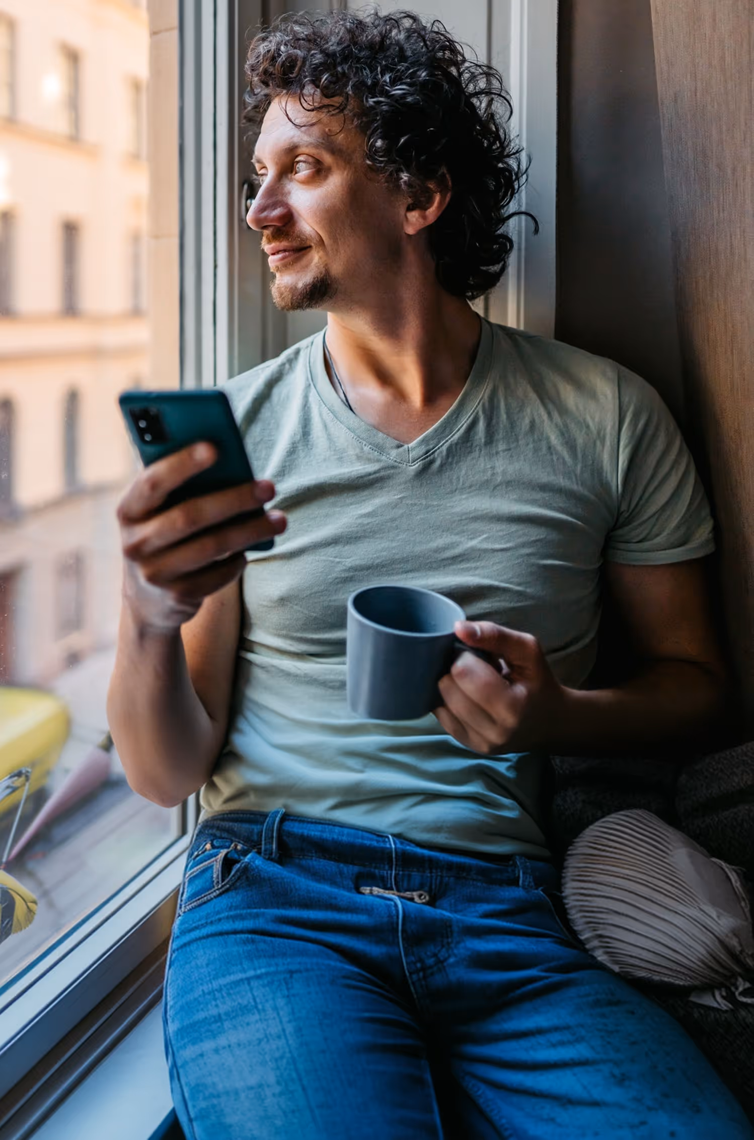 Mand med krøllet hår sidder ved vindue med telefon i ene hånd og koffeekop i den anden.