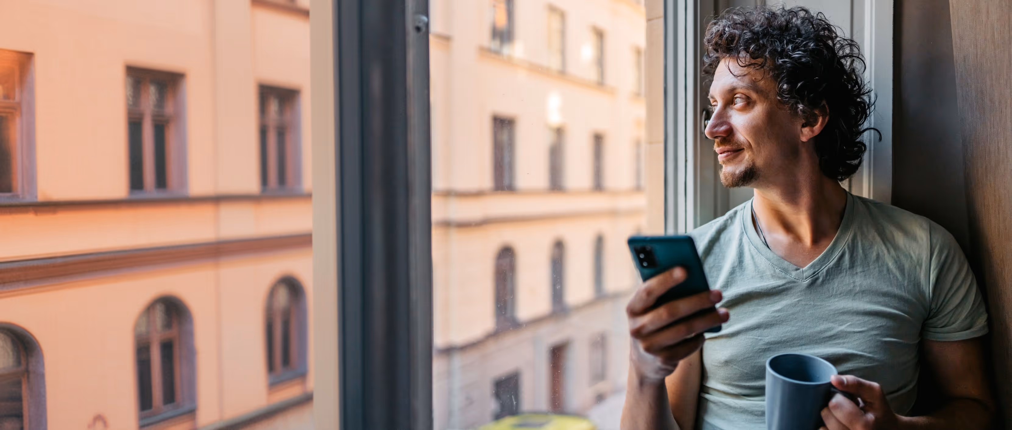 Mand med krøllet hår ser ud af vinduet med en telefon i den ene hånd og en kop i den anden.