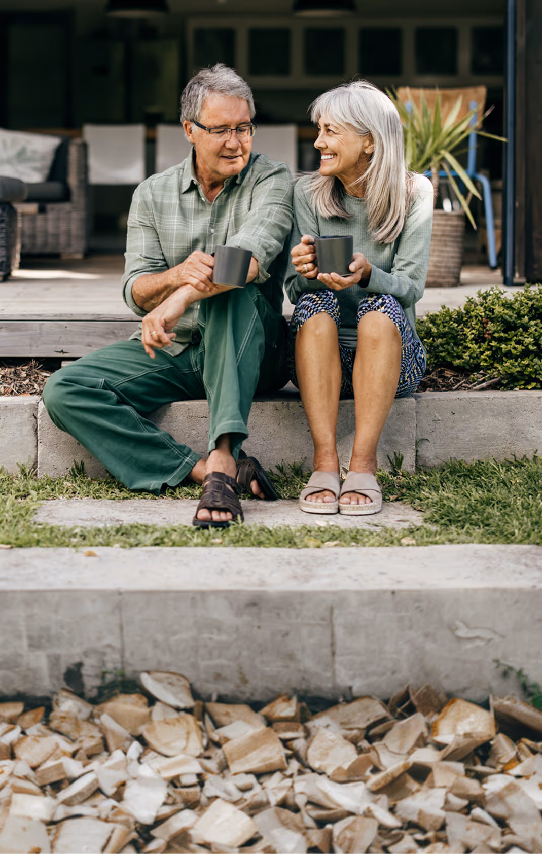 Smiling elderly couple sitting outside on a porch step holding mugs, enjoying a moment together.