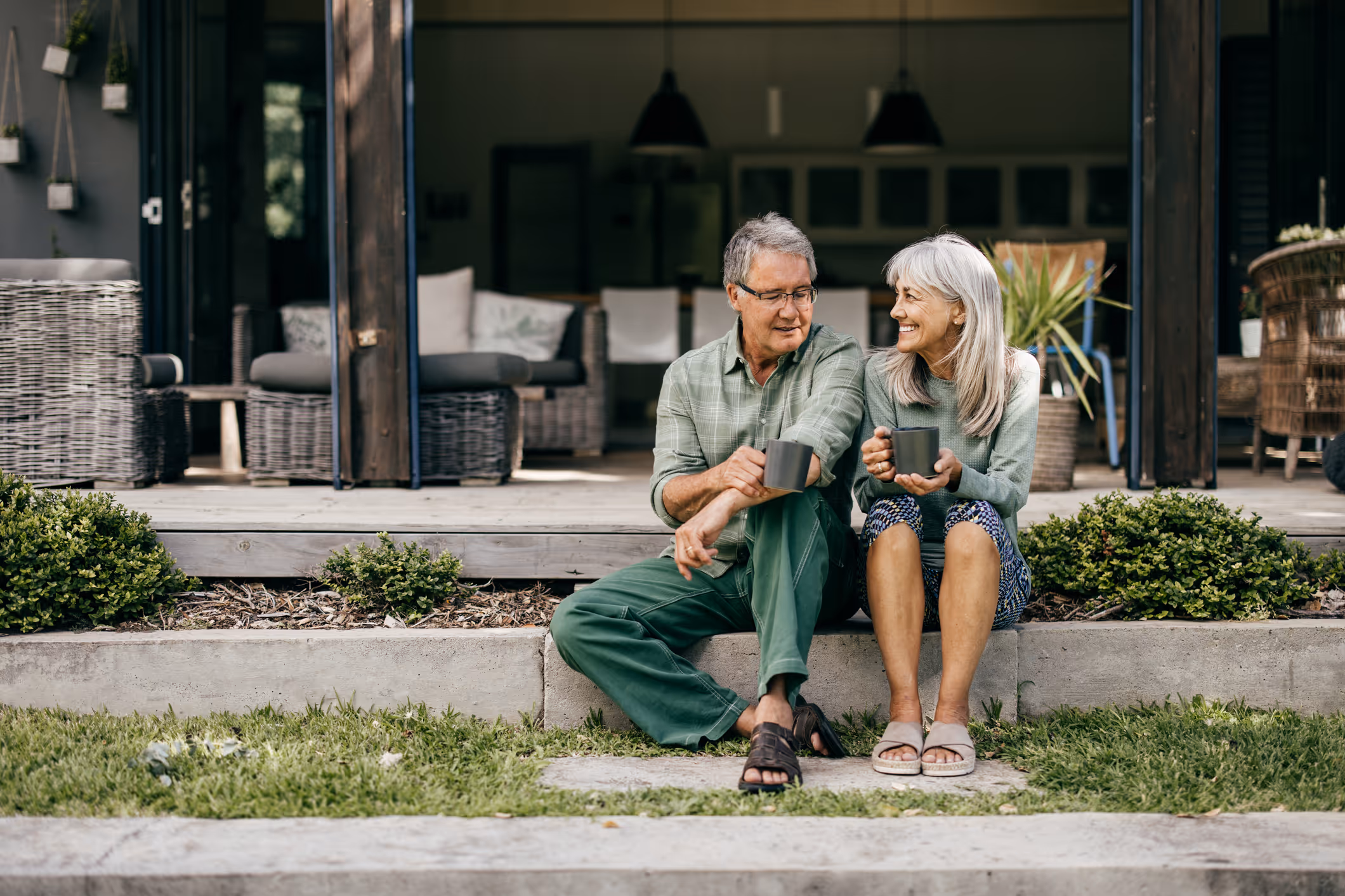 Smiling older couple sitting outside a modern home, holding mugs and enjoying each other's company.