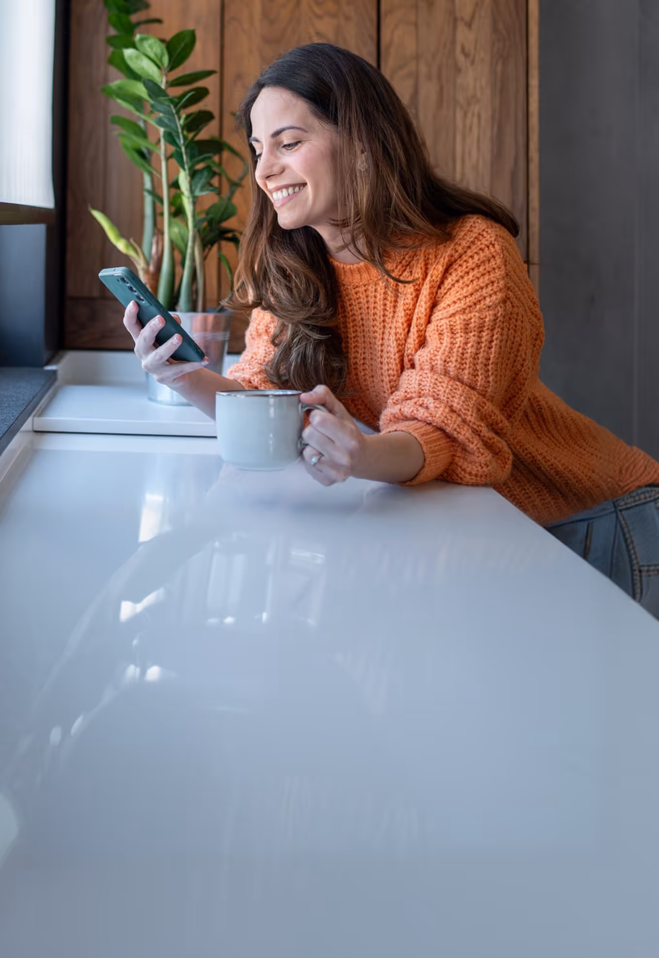 Kvinde i orange trøje smiler, mens hun sidder ved bordet med en kop og tjekker sin telefon.