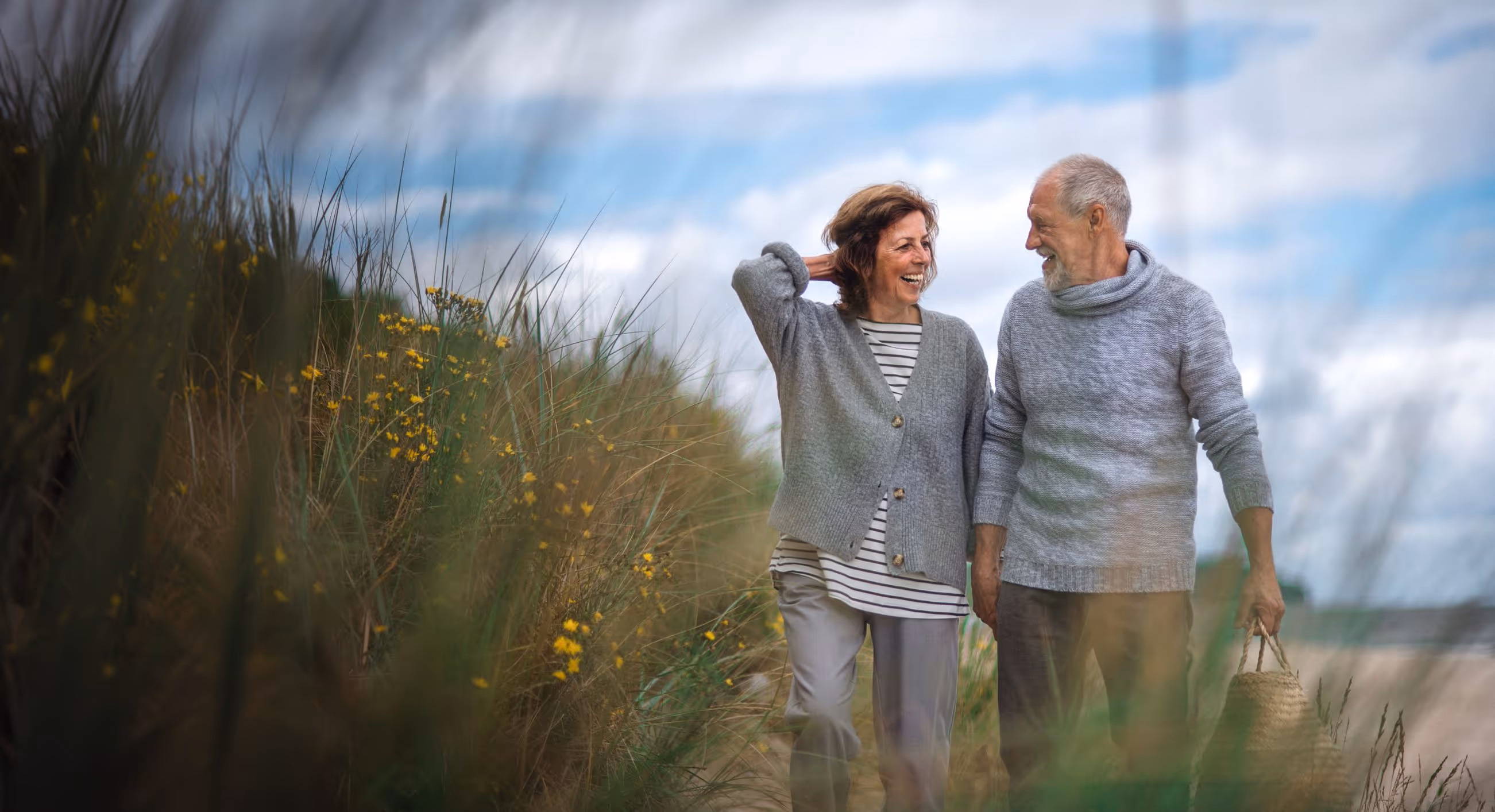 Smiling elderly couple walking outdoors near tall grass and yellow flowers on a cloudy day.