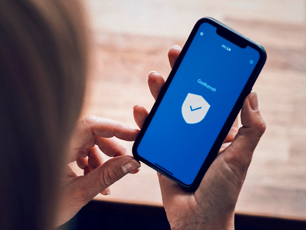 Person holder en smartphone med en blå skærm, der viser et hvidt skjold med et flueben og teksten Godkendt.