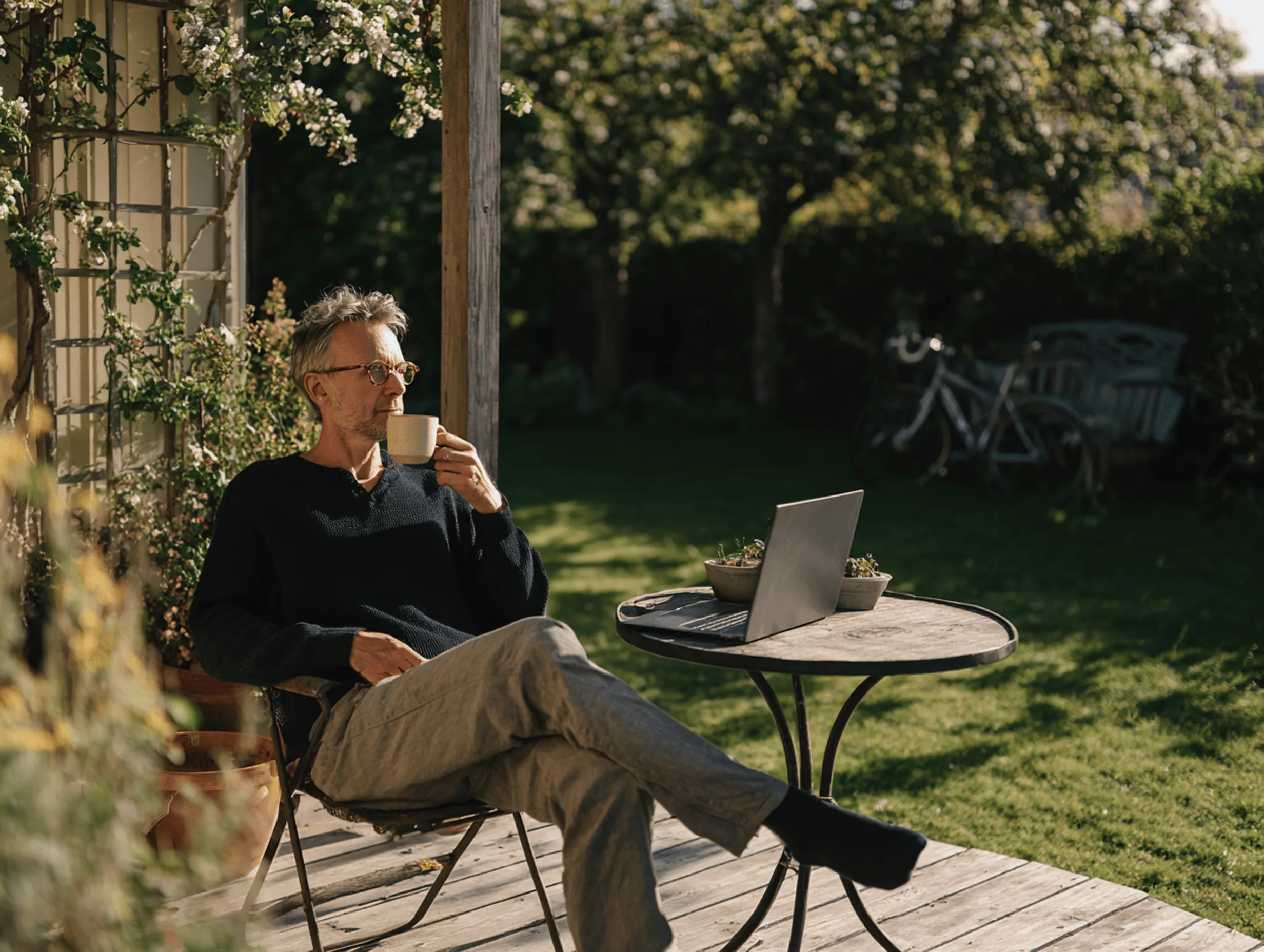Mand sidder på terrasse med kaffekop og laptop, omgivet af blomster og have i sollys.